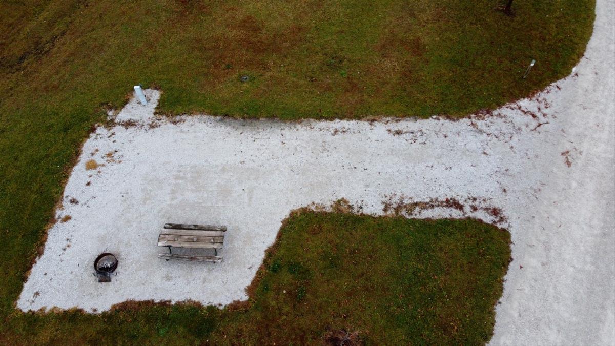 Aerial view of empty camp site with limestone pad, picnic table, fire ring, and trees. 