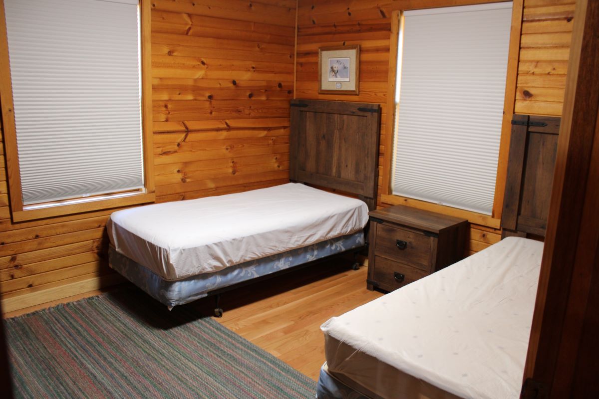Two twin beds with a nightstand in between and 2 windows with shades drawn 