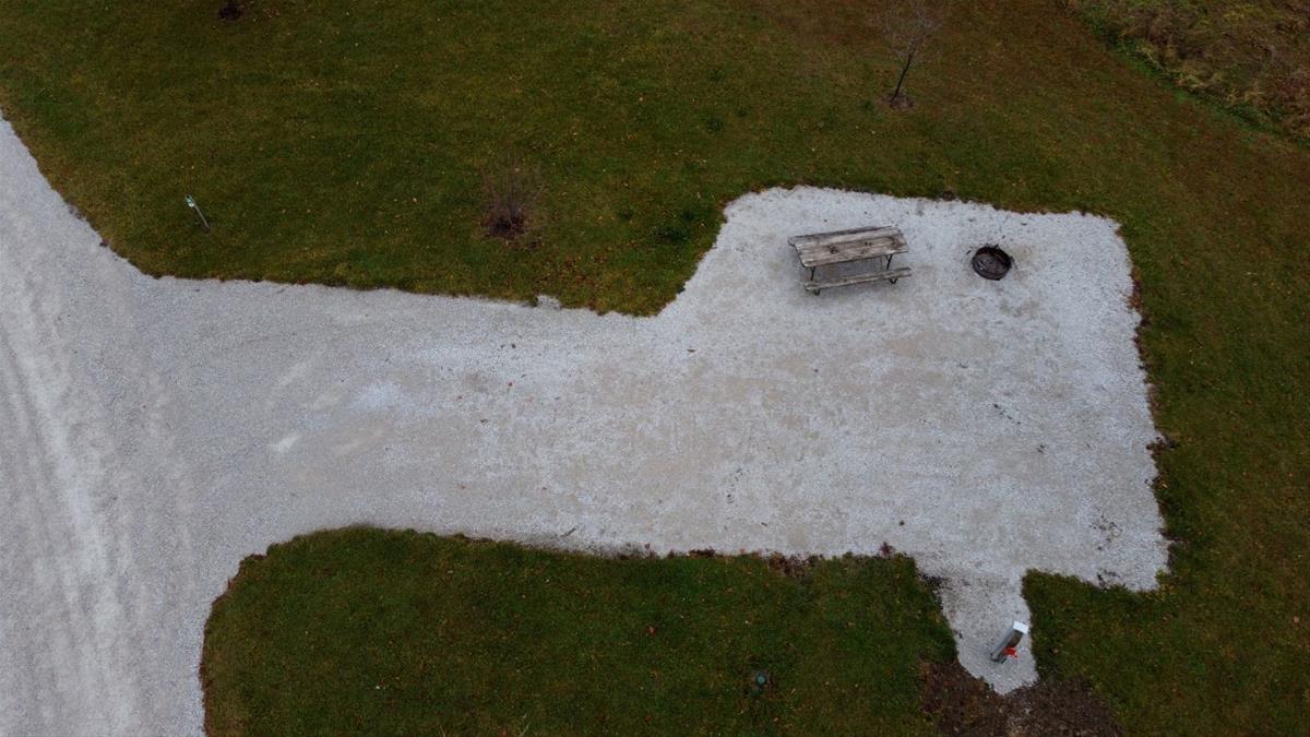 Aerial view of empty camp site with limestone pad, picnic table, fire ring, and trees. 