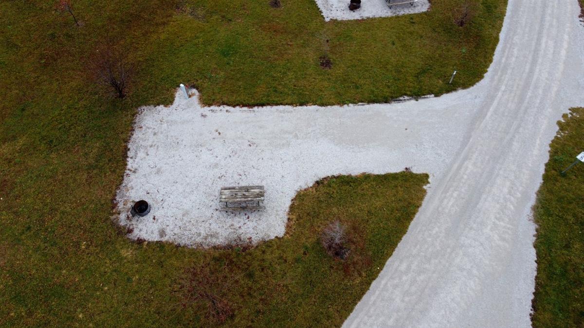 Aerial view of empty camp site with limestone pad, picnic table, fire ring, and trees. 