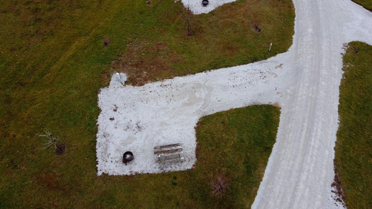 Aerial view of empty camp site with limestone pad, picnic table, fire ring, and trees. 