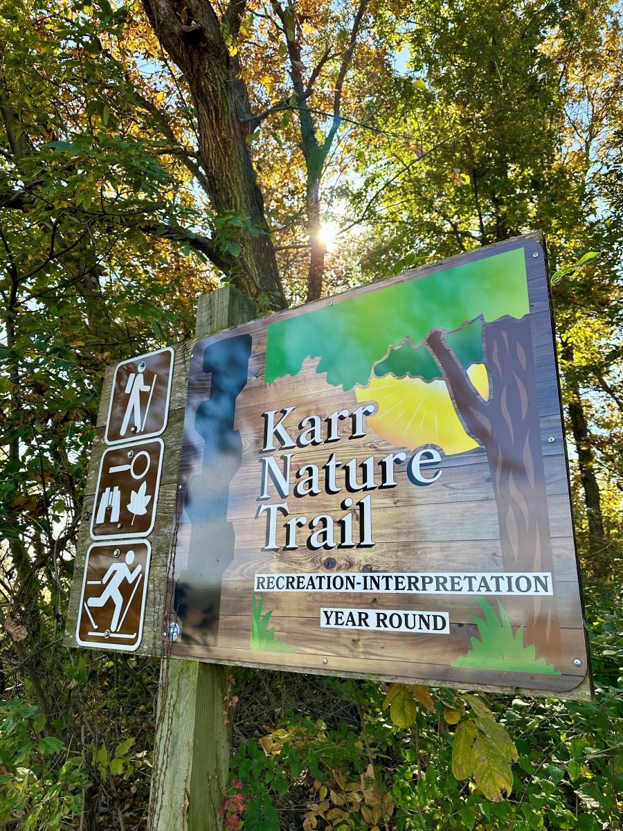 Trailhead sign for the Karr Nature Trail with sun shining through tree canopy in back 