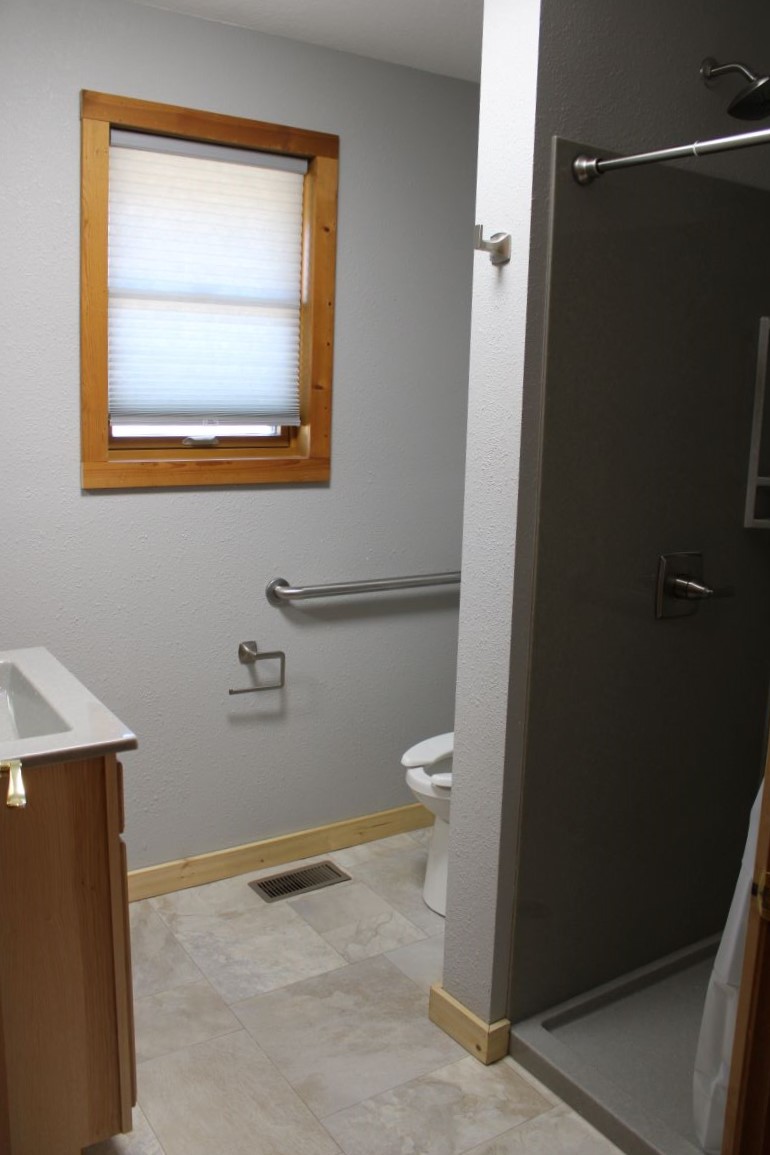 Bathroom with walk in shower and sink, window with shade drawn and hand rail near toilet