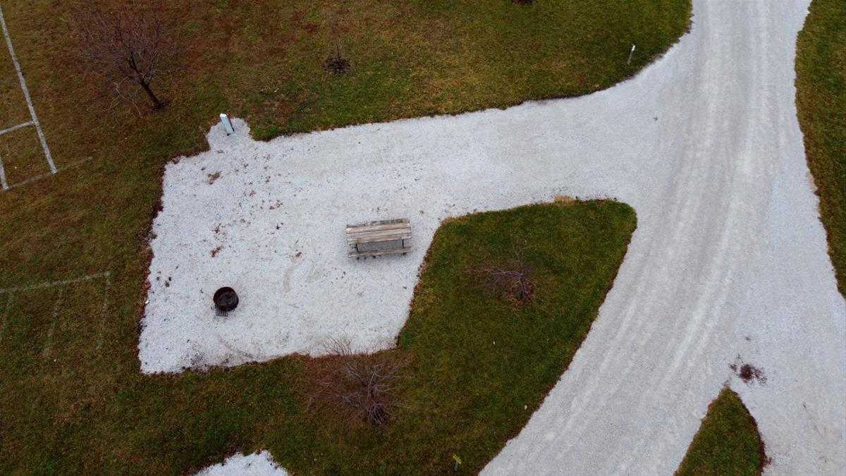 Aerial view of empty camp site with limestone pad, picnic table, fire ring, and trees. 