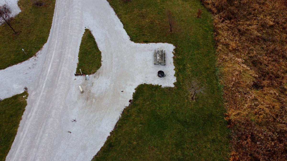 Aerial view of empty camp site with limestone pad, picnic table, fire ring, and trees. 
