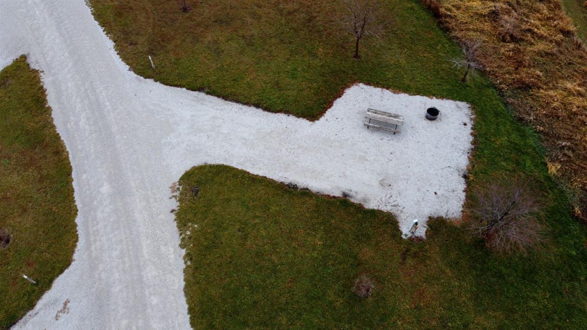 Aerial view of empty camp site with limestone pad, picnic table, fire ring, and trees. 