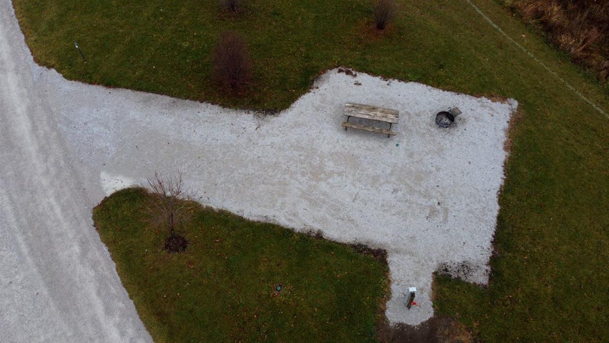 Aerial view of empty camp site with limestone pad, picnic table, fire ring, and trees. 