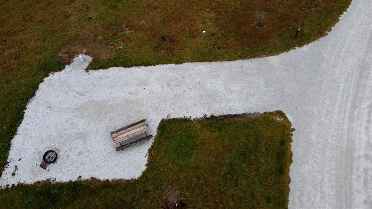Aerial view of empty camp site with limestone pad, picnic table, fire ring, and trees. 